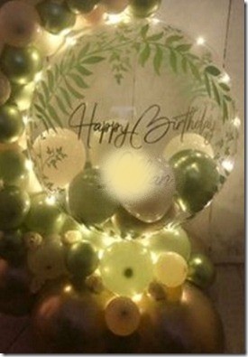 Happy birthday print on the transparent balloon with small White and green balloons stuffed inside and outside like a balloon garland decorated with foliage white flowers and led lights
